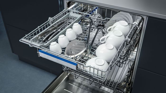 Un panier de lave-vaisselle chargé avec des assiettes blanches, des tasses, des couverts et une passoire, prêt pour le nettoyage dans une cuisine moderne.