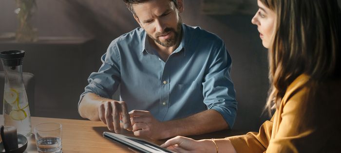 Siniseen paitaan pukeutunut mies osoittaa puupöydällä olevaa tablettia. 