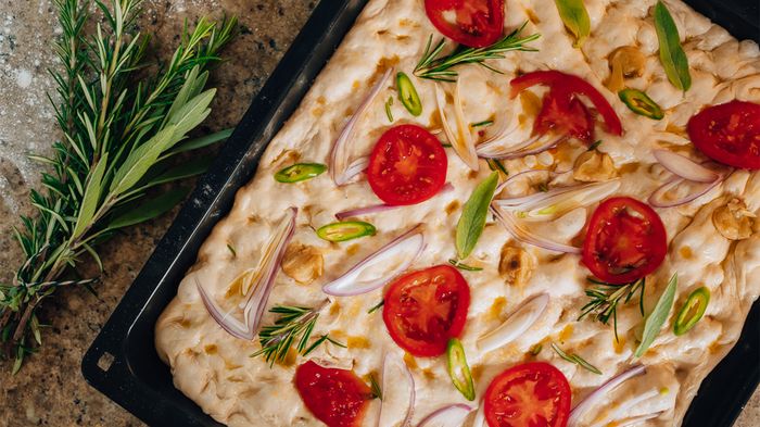 Vista dall&rsquo;alto di una focaccia in teglia con pomodorini, cipolla a fettine ed erbe aromatiche