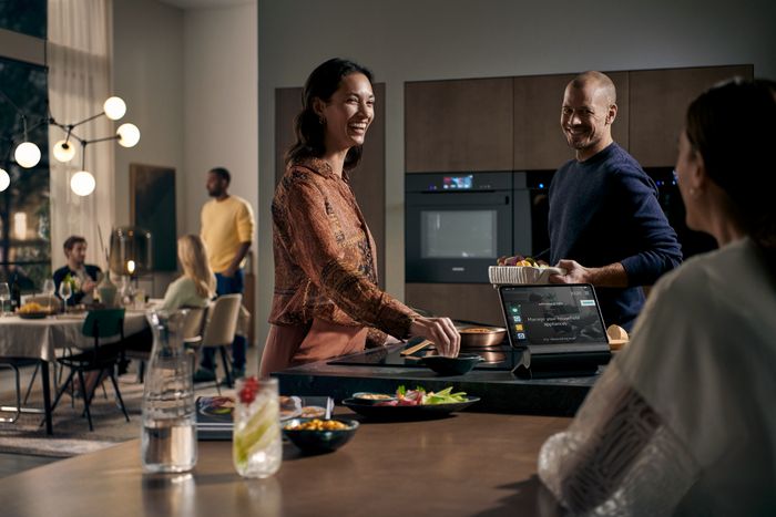 En moderne k&oslash;kkenscene med venner, der nyder et m&aring;ltid, hvor en person serverer mad, og der er en touchscreen-enhed p&aring; bordpladen.