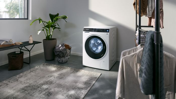 Lavadora Siemens de carga frontal en cuarto de lavado moderno, decorado con plantas, ropa colgada y luz natural entrando por la ventana.