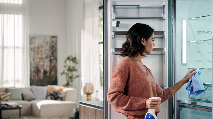 Qualcuno pulisce l'interno di un frigorifero aperto con un panno blu in un soggiorno luminoso e moderno.