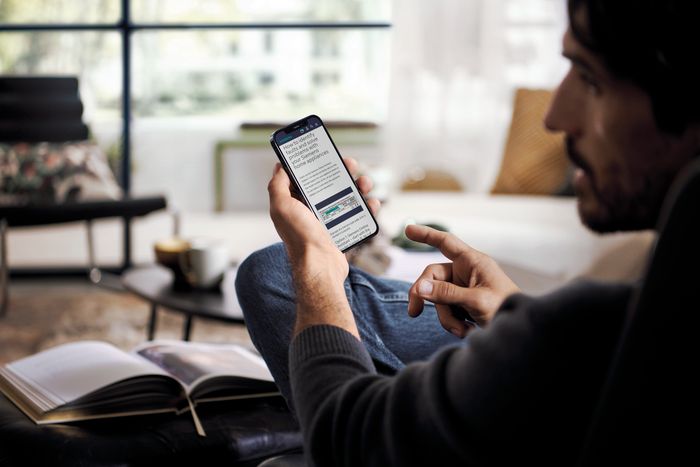 Osoba siedząca swobodnie w przytulnym pokoju, trzymająca smartfon i przewijająca coś, w pobliżu ma książkę i filiżankę kawy.