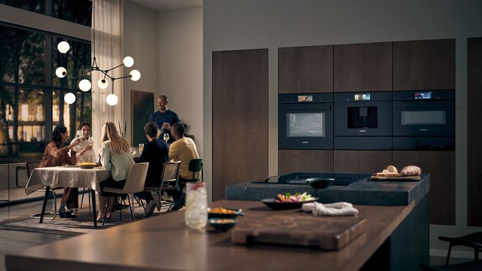 Moderne Küche mit Essbereich, Menschen sitzen am Tisch, Küchengeräte und dekorative Beleuchtung. 