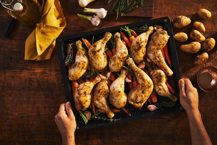 Mani che tengono una teglia con cosce di pollo e verdure arrosto su tavolo in legno