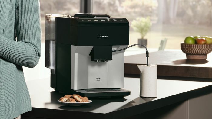 Siemens aparat za kafu na kuhinjskoj radnoj površini sa posudom za mleko i kolačićima pored njega