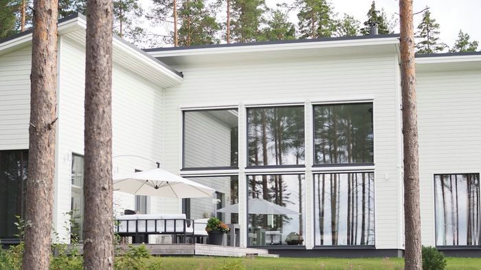 A modern white house with large windows and a patio, surrounded by lush greenery.