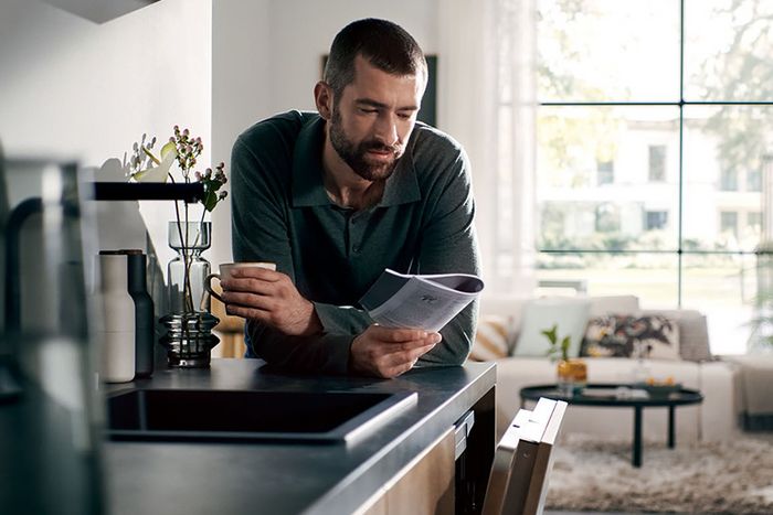 Un hombre lee un manual de instrucciones en la cocina mientras toma una taza de café. Se apoya en la encimera, donde hay una placa de cocina y algunos objetos decorativos. Al fondo se ve el salón.