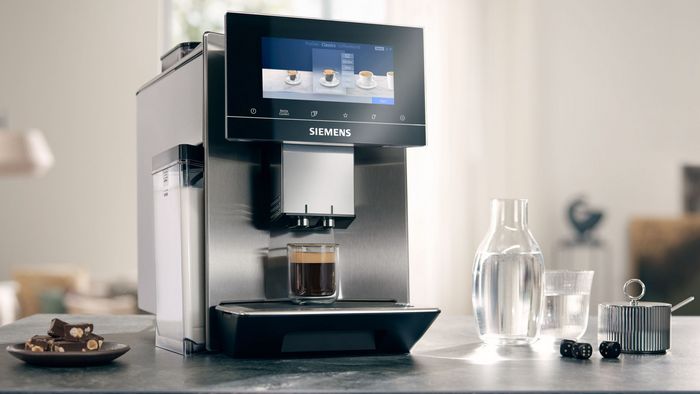 La machine à café entièrement automatique EQ900 est posée sur un plan de travail en granit, à côté duquel se trouvent une carafe d'eau avec un verre, des pâtisseries sur une petite assiette et un sucrier.