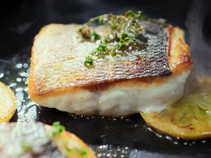 A close-up of a perfectly seared fish fillet with crispy skin and fresh herbs, cooking in a pan with lemon slices, emitting steam.
