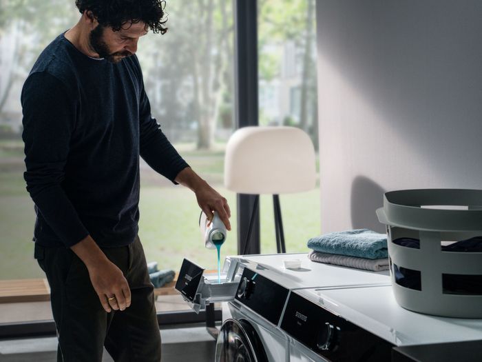 Une personne verse du détergent dans une machine à laver, avec une lampe moderne et un panier de linge à l'arrière-plan.