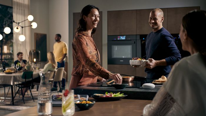 Moderní kuchyně, žena připravující jídlo, muž držící talíř, chytrý reproduktor na lince u stolu během setkání.