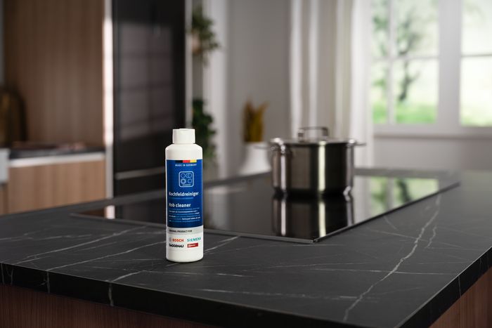 Flacone di detergente Cleaning&Care Siemens su un piano cucina in marmo scuro, con piano cottura a induzione e pentola sullo sfondo
