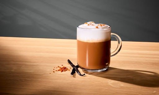 Een latte met dikke melkschuimlaag in een glas, ernaast liggen wat suiker en stokjes vanille.