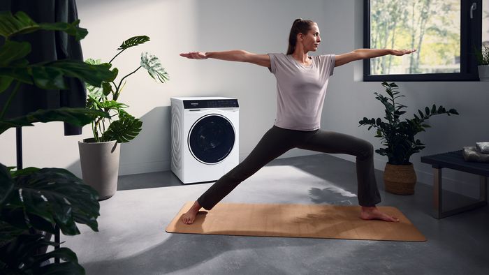 Eine Frau steht in der Waschküche vor einem Siemens iQ700 Waschtrockner. Sie praktiziert die Yoga-Übung der Krieger.