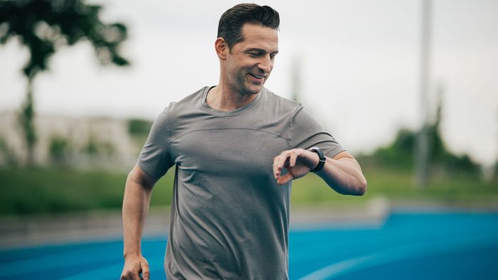 Man in Outdoorbekleidung auf einer Tartanbahn blickt auf seine Sportuhr.