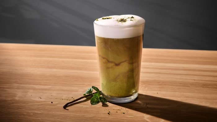 Een glas met groene matcha-koffie met schuimlaag, om het glas heen liggen een kaneelstokje, een blaadje en wat matcha-poeder.