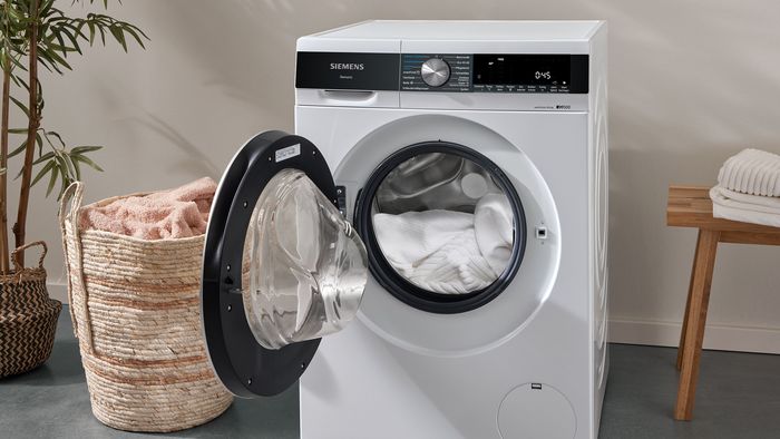 Uma m&aacute;quina de lavar roupa aberta, cheia de roupa branca, est&aacute; ao lado de uma cesta de roupa de grama.