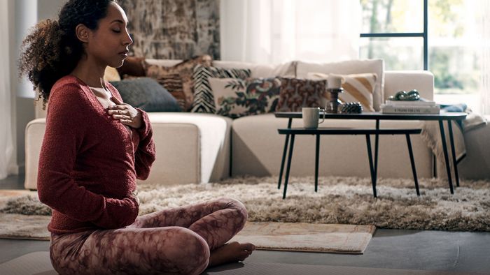 Uma mulher senta-se numa esteira de yoga numa sala de estar a fazer exerc&iacute;cios de respira&ccedil;&atilde;o.