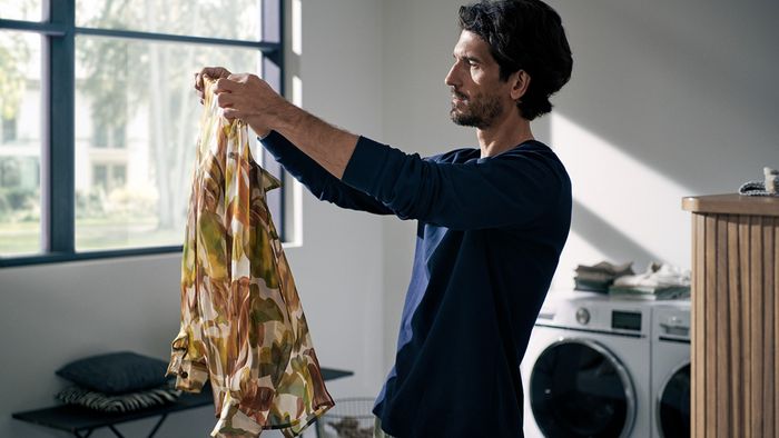 Ein Mann h&auml;lt einen gemusterten Stoff ans Fensterlicht in einem hellen Waschraum mit einer Waschmaschine und einem Trockner im Hintergrund.