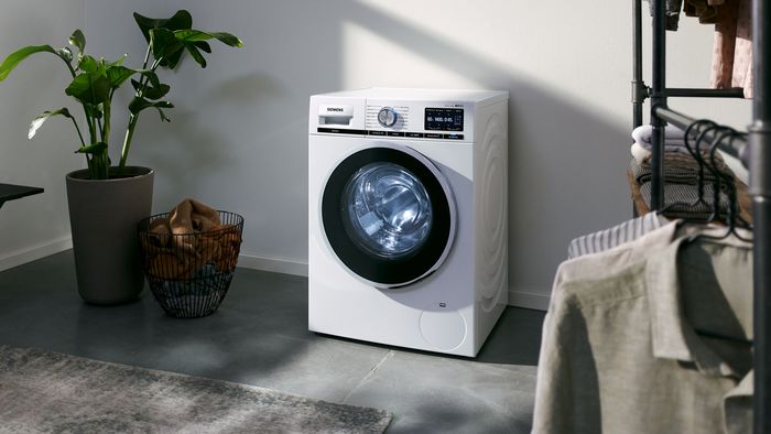 Eine Waschmaschine steht in einem hell erleuchteten Raum. Im Vordergrund hängt frische Wäsche auf einem Seil. Neben der Maschine befindet sich eine hohe Gewächshauspflanze.