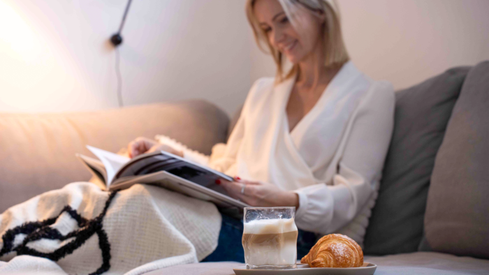 Une femme lit un magazine, install&eacute;e sous un plaid dans un fauteuil. Au premier plan, un petit caf&eacute; latte et un croissant sont dispos&eacute;s sur une  assiette.