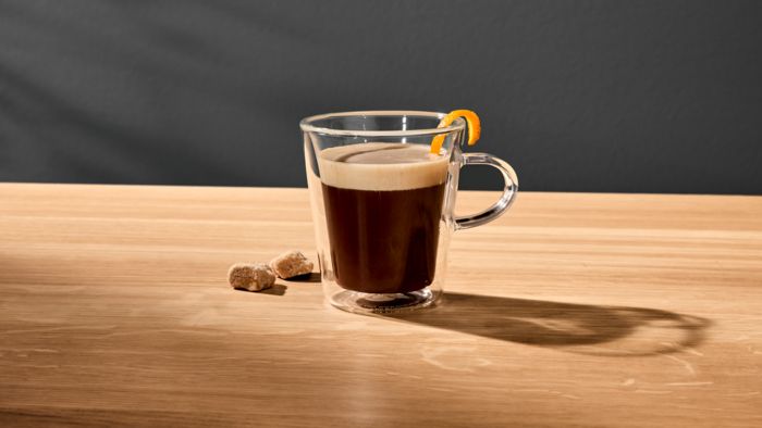 Koffie in een glas met een sinaasappelschil over de rand en een paar suikerklontjes op de achtergrond.