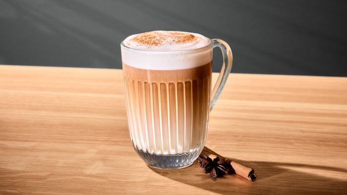 Koffie in geribbeld glas met melkschuim en kaneel, op de achtergrond een kaneelstokje en steranijs.