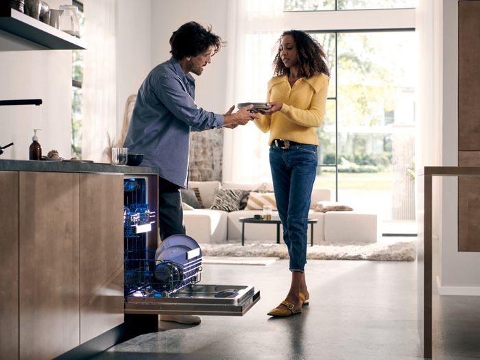 Coppia in cucina: uomo porge un piatto a una donna mentre la lavastoviglie incassata &egrave; aperta con luce interna blu