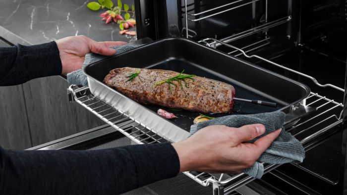 Eine Person nimmt eine Pfanne mit einem gewürzten Braten aus dem Ofen und verwendet dabei ein Küchentuch und ein Fleischthermometer.