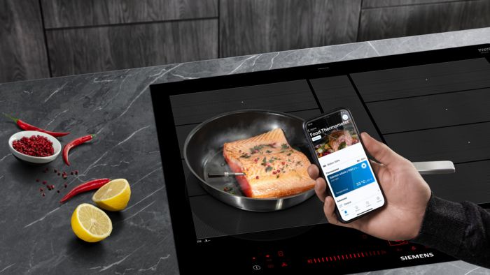 Eine Person überwacht das Kochen von Lachs auf einem intelligenten Induktionskochfeld mit einem Smartphone