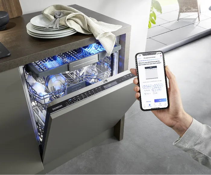 יד של אדם אוחז מכשיר סלולרי שמתחבר באפליקציה למדיח הכלים של סימנס שמופיע ברקע