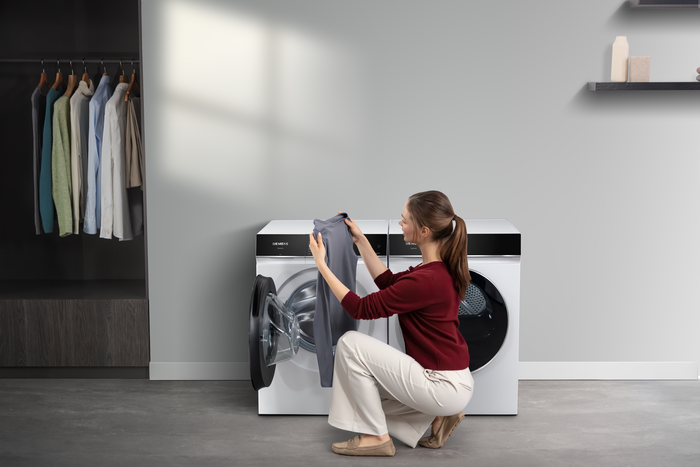 Eine Frau l&auml;dt W&auml;sche in eine wei&szlig;e Siemens Frontlader‑Waschmaschine in einem modernen Ankleidezimmer. 