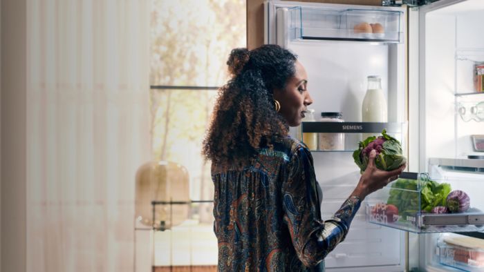תמונה של אישה עם שיער תלתל רואה את תוכן המקרר, מחזיקה סלט ומוצרי חלב. המקרר הוא של Siemens ומואר באור טבעי שנכנס מהחלון.