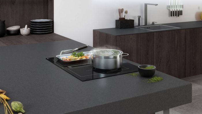 Placa FlexInducción de 70 cm con extractor incorporado en cocina moderna con encimeras y muebles oscuros, con ingredientes y utensilios alrededor.