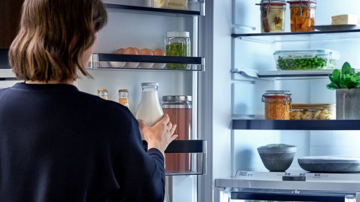 Una mujer de espaldas coge una botella de leche de la puerta de un frigorífico Siemens, abierto y bien organizado.