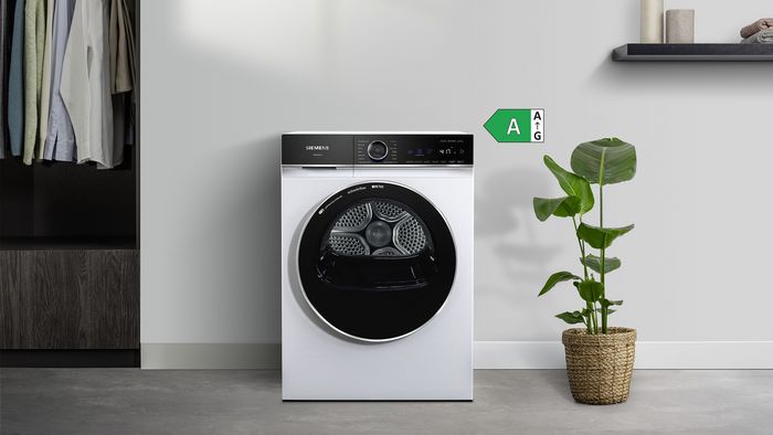  En moderne Siemens-t&oslash;rretumbler i hvid st&aring;r ved siden af en potteplante, med p&aelig;nt oph&aelig;ngt t&oslash;j i baggrunden. Energim&aelig;rkningen er synlig.