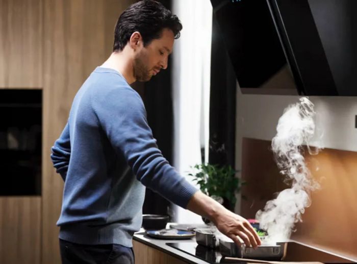 A man is preparing food in a kitchen, with steam billowing from the stove, indicating active cooking.  