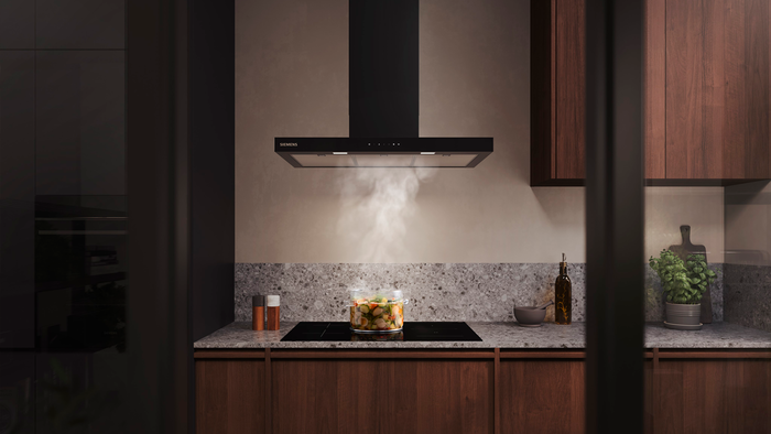 In einer modernen Küche wird Gemüse in einem Glaskochtopf auf einem Induktionskochfeld gedämpft, darüber eine Dunstabzugshaube.