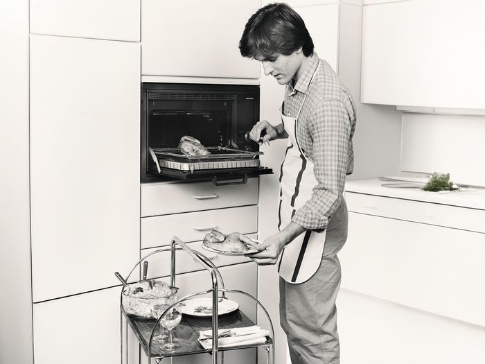 Ein Mann mit einer Sch&uuml;rze zieht ein Backblech mit Brath&auml;hnchen aus dem Backofen. Ein Servierwagen mit Tellern und Gl&auml;sern steht in Reichweite.