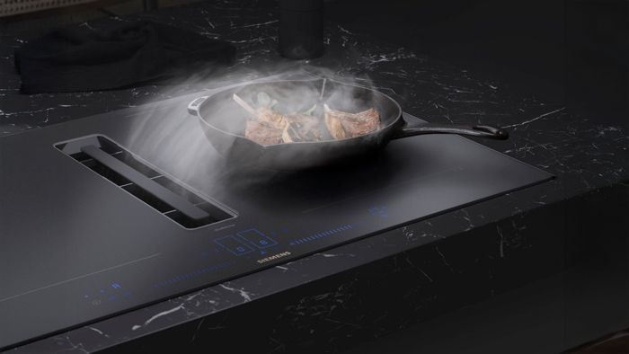 Siemens matzwarte inductiekookplaat met een pan met lamskoteletten, stoom die oprijst, geplaatst tegen een  zwart marmeren oppervlak. 