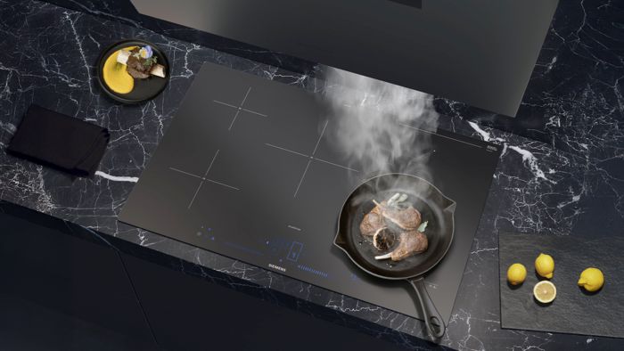 Matzwarte Siemens inductiekookplaat met een pan met gebakken lam, omgeven door verse citroenen op een donker marmeren oppervlak.