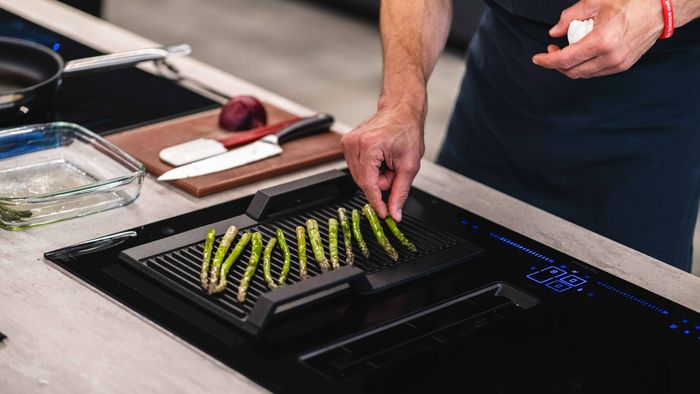 A man grills asparagus on a venting hob, showcasing vibrant green spears cooking over an open flame.  