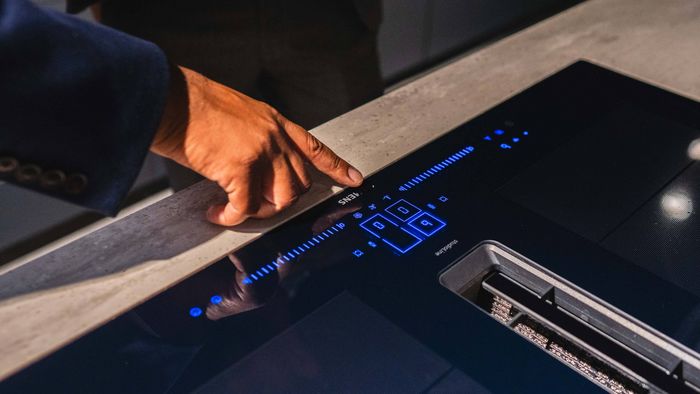 A person touches a blue light on a countertop, indicating the operation of a venting hob. 