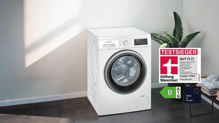 „Siemens Waschmaschine WU14UT71 mit Stiftung Warentest Testsieger-Siegel GUT (1,7), Energielabel B und Hinweis ‚Made in Europe‘ in modernem Raum mit Pflanze und gefalteter Wäsche.