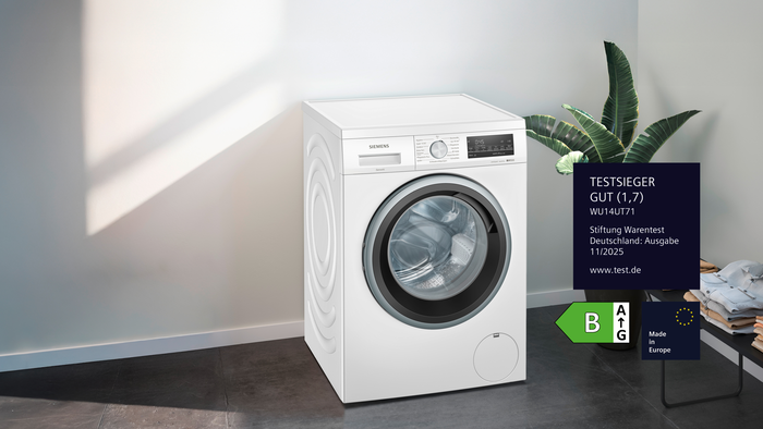 &bdquo;Siemens Waschmaschine WU14UT71 mit Stiftung Warentest Deutschland GUT (1,7), Energielabel B und Hinweis &sbquo;Made in Europe&lsquo; in modernem Raum mit Pflanze und gefalteter W&auml;sche.