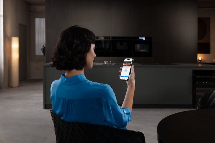 A woman uses a smartphone app to control Siemens kitchen appliances, including an oven, coffee machine, and microwave, in a modern, dark-toned kitchen.