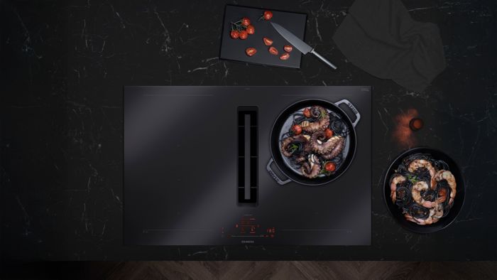 &nbsp;Een slank zwart keukenblad met een pan zeevruchtenpasta, verse tomaten, een mes en een schaal garnalen op een marmeren oppervlak.