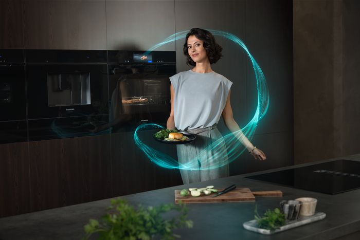 A woman holds a plate of cooked salmon and broccoli, surrounded by glowing blue digital lines, in a modern kitchen with integrated Siemens appliances.