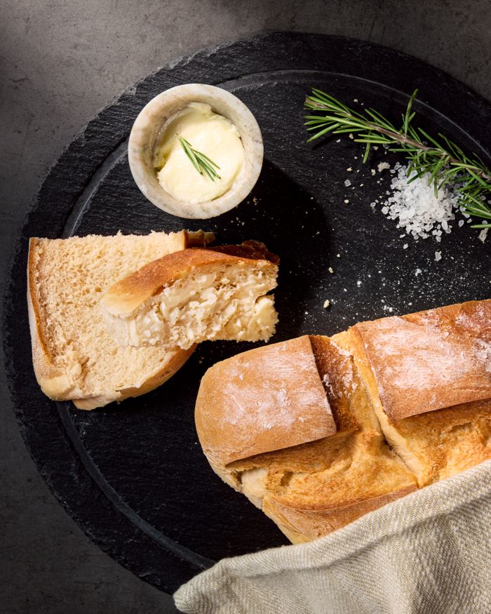 Freshly baked bread, a bowl of butter with rosemary, and coarse salt on a dark slate serving board.